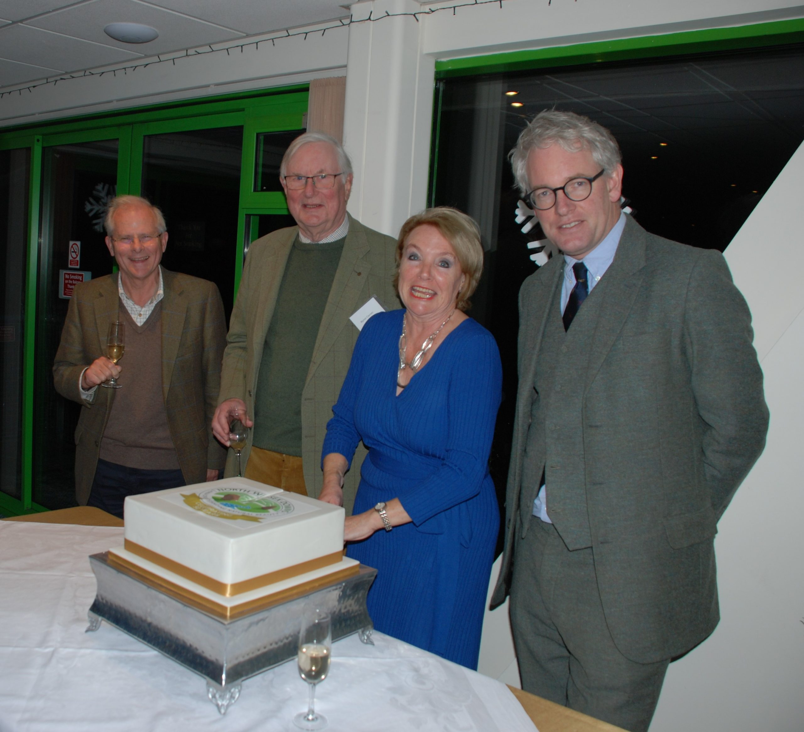 A woman is cutting a celebratory cake while three men stand beside her and look at the camera