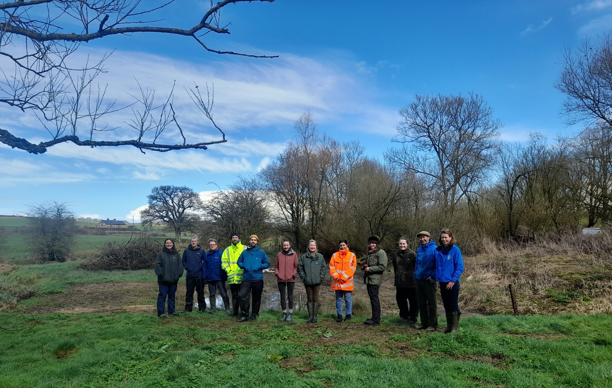 12 people are standing in a line in a field in front of a pond. 