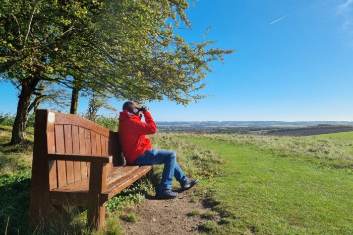 A man in a red jacket is sitting on a wooden bench looking through binoculars. There is a tree behind him and views beyond and in front of him